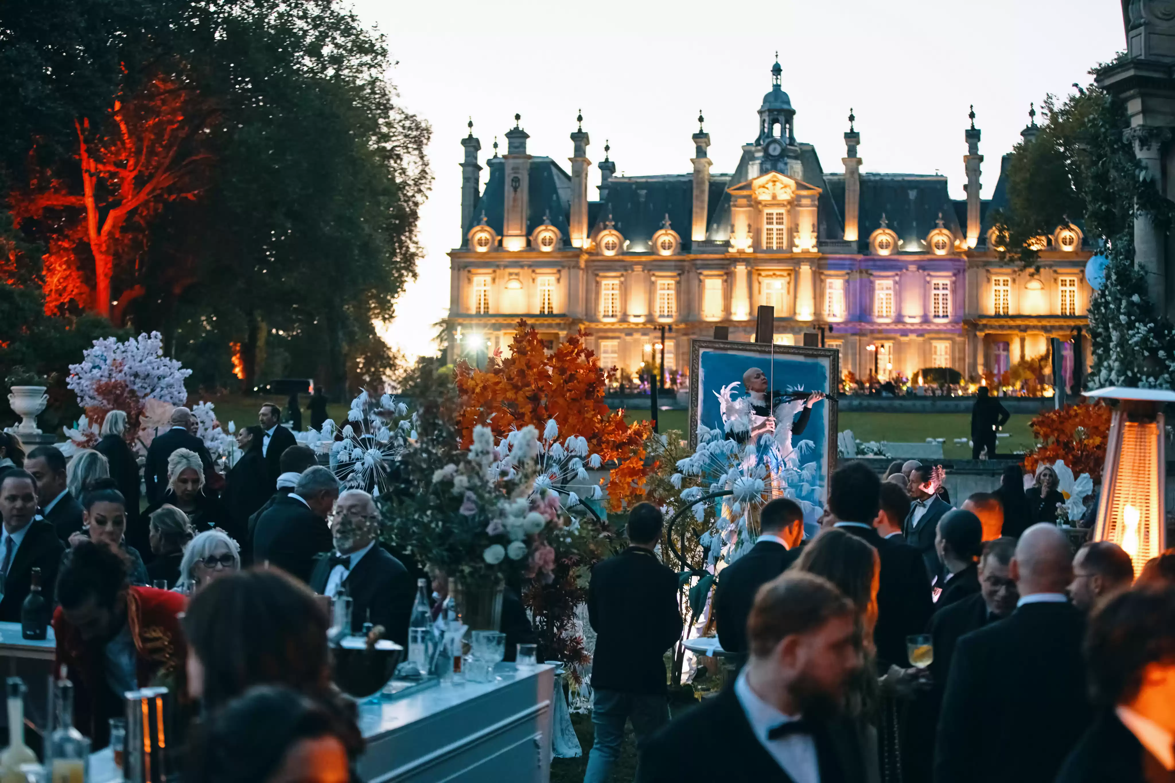 Réception de mariage grandiose en extérieur à la tombée de la nuit, avec le château illuminé en arrière-plan, de nombreux invités, un bar à cocktails et une animation ar