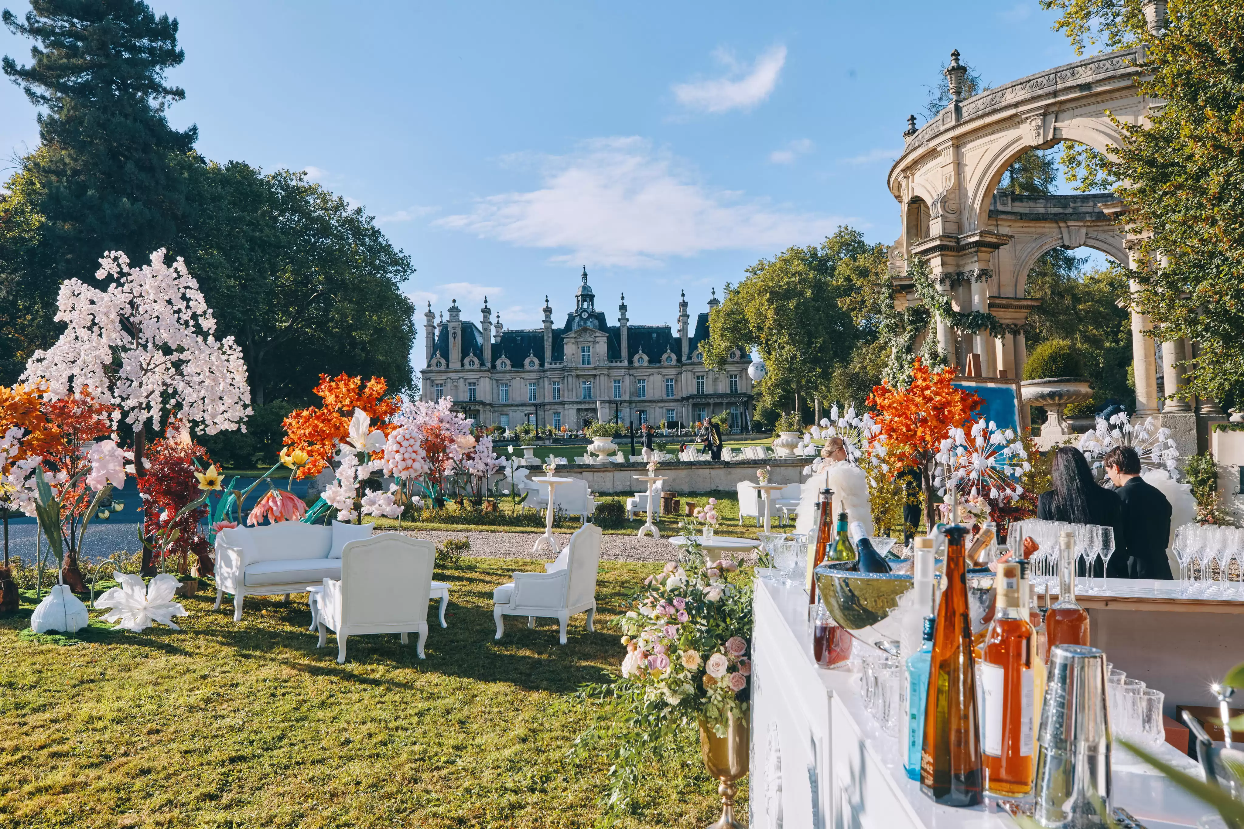 Ambiance de cocktail grandiose sur la pelouse centrale à l'entrée du domaine, avec des salons blancs élégants, une décoration florale colorée et le château majestueux en arriè