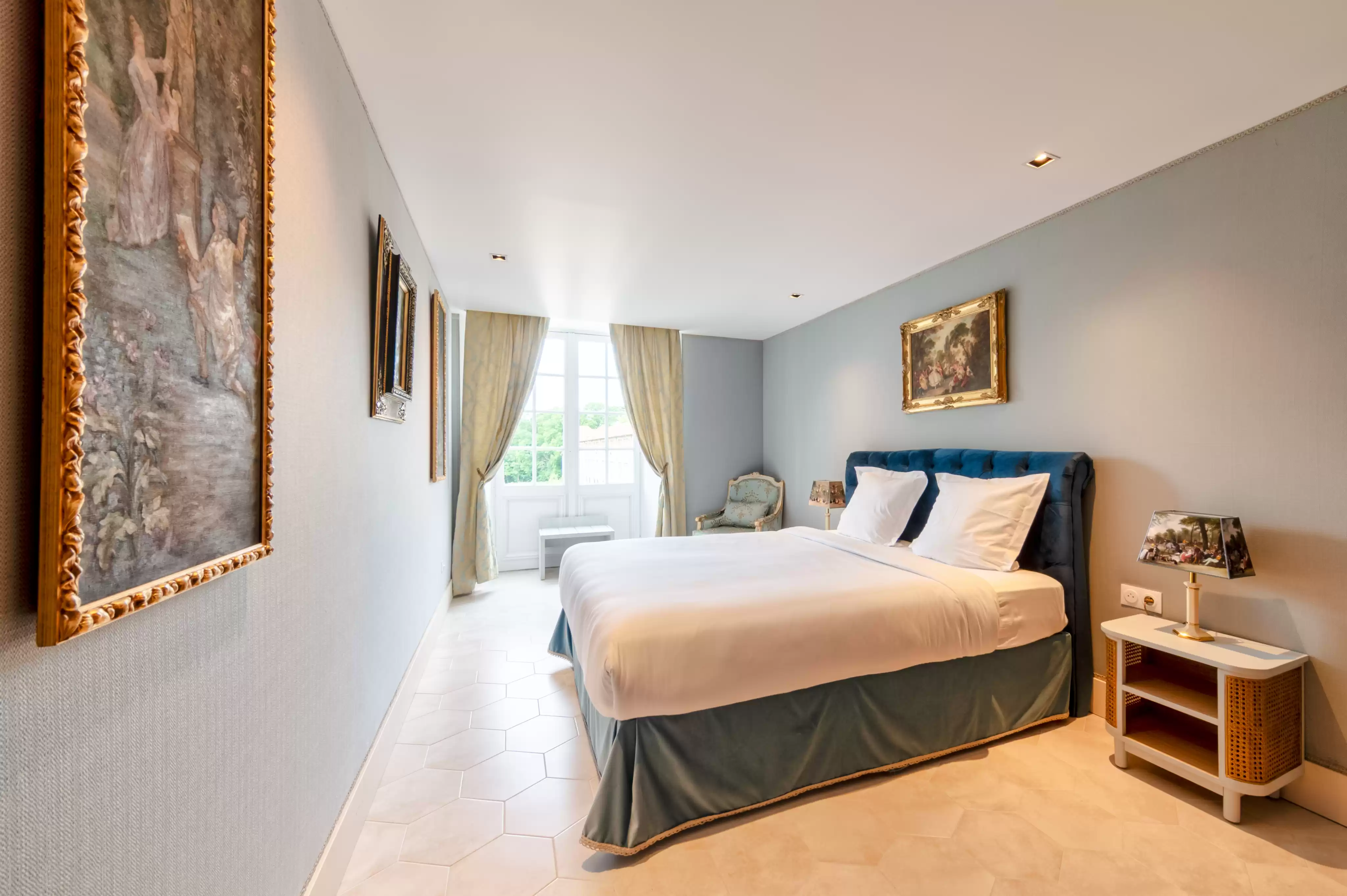 Chambre raffinée et lumineuse avec vue sur le parc, décorée de murs gris-bleu doux, d'un lit avec tête de lit en velours bleu, et de multiples tableaux anciens encad