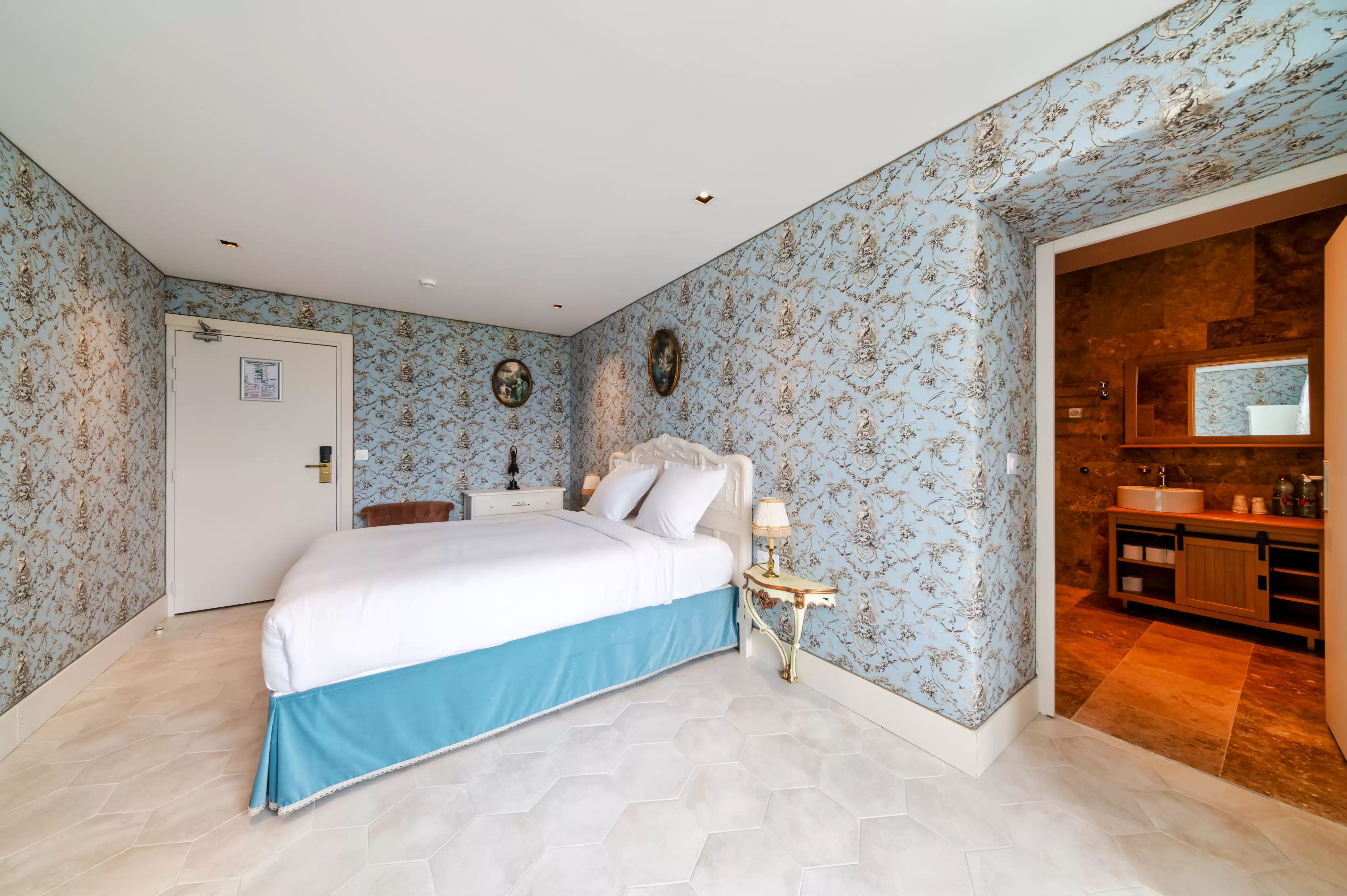 Chambre authentique et chaleureuse avec un papier peint bleu à motifs élégants, un lit confortable avec une jupe de lit bleue, et un aperçu de la salle de bain attena