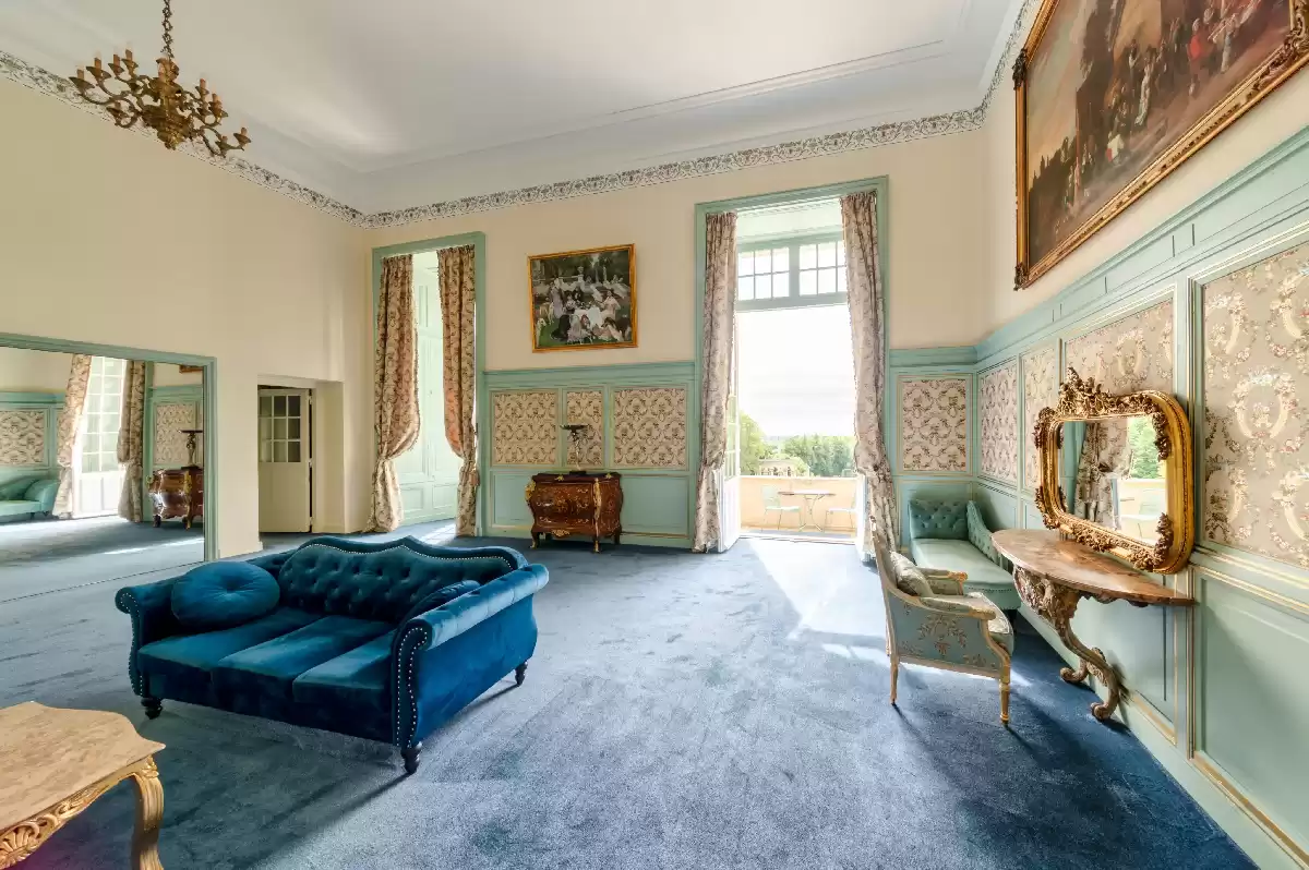 Vue d'ensemble du spacieux et lumineux salon de préparation de la mariée, doté d'élégantes boiseries bleu pastel et dorées, d'une moquette bleue assortie, d'un grand canapé en velours bleu et ouvrant sur un balcon av