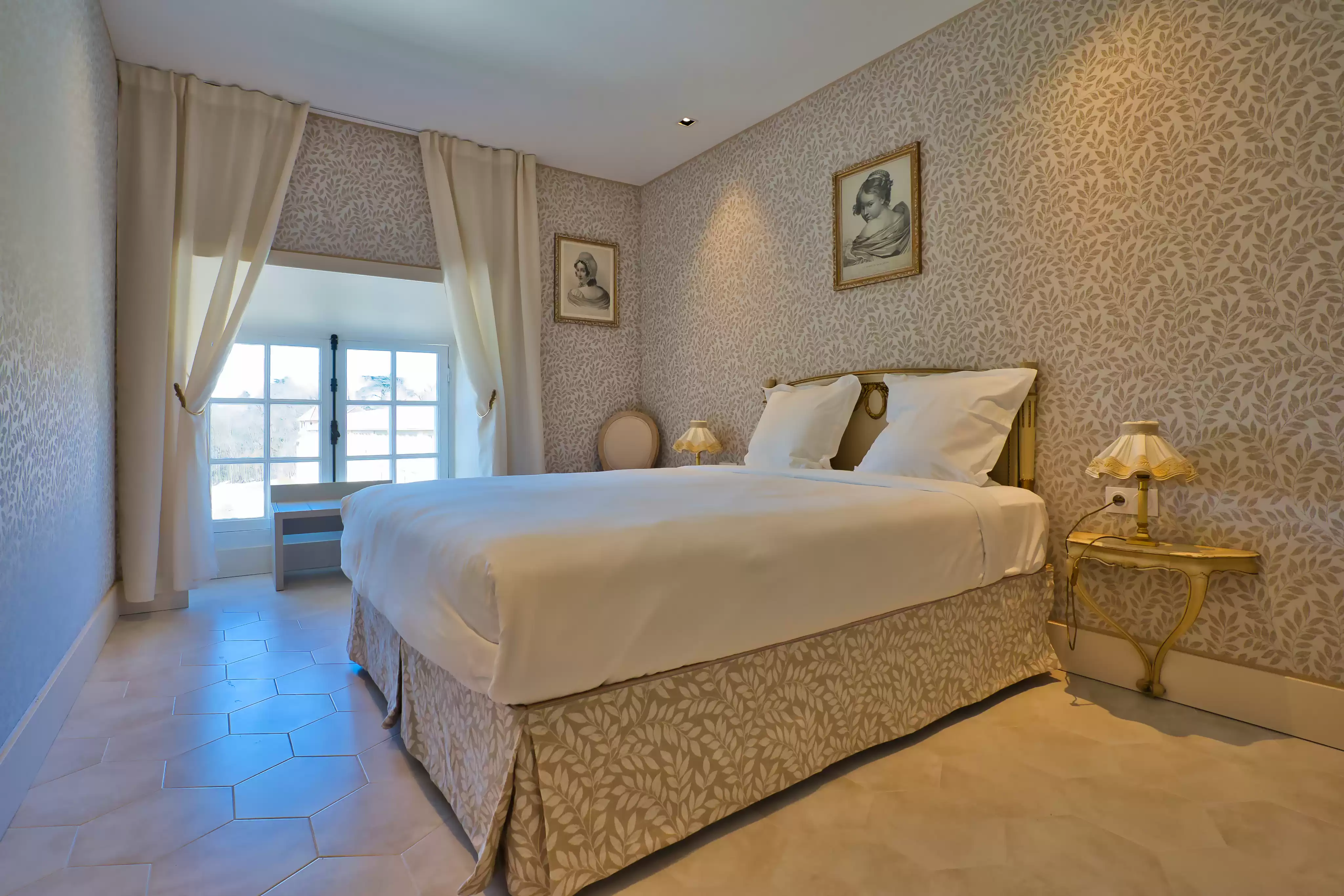 Élégante chambre de la suite familiale décorée dans un esprit classique et raffiné, avec un papier peint à motifs délicats, un grand lit double et une table de chevet