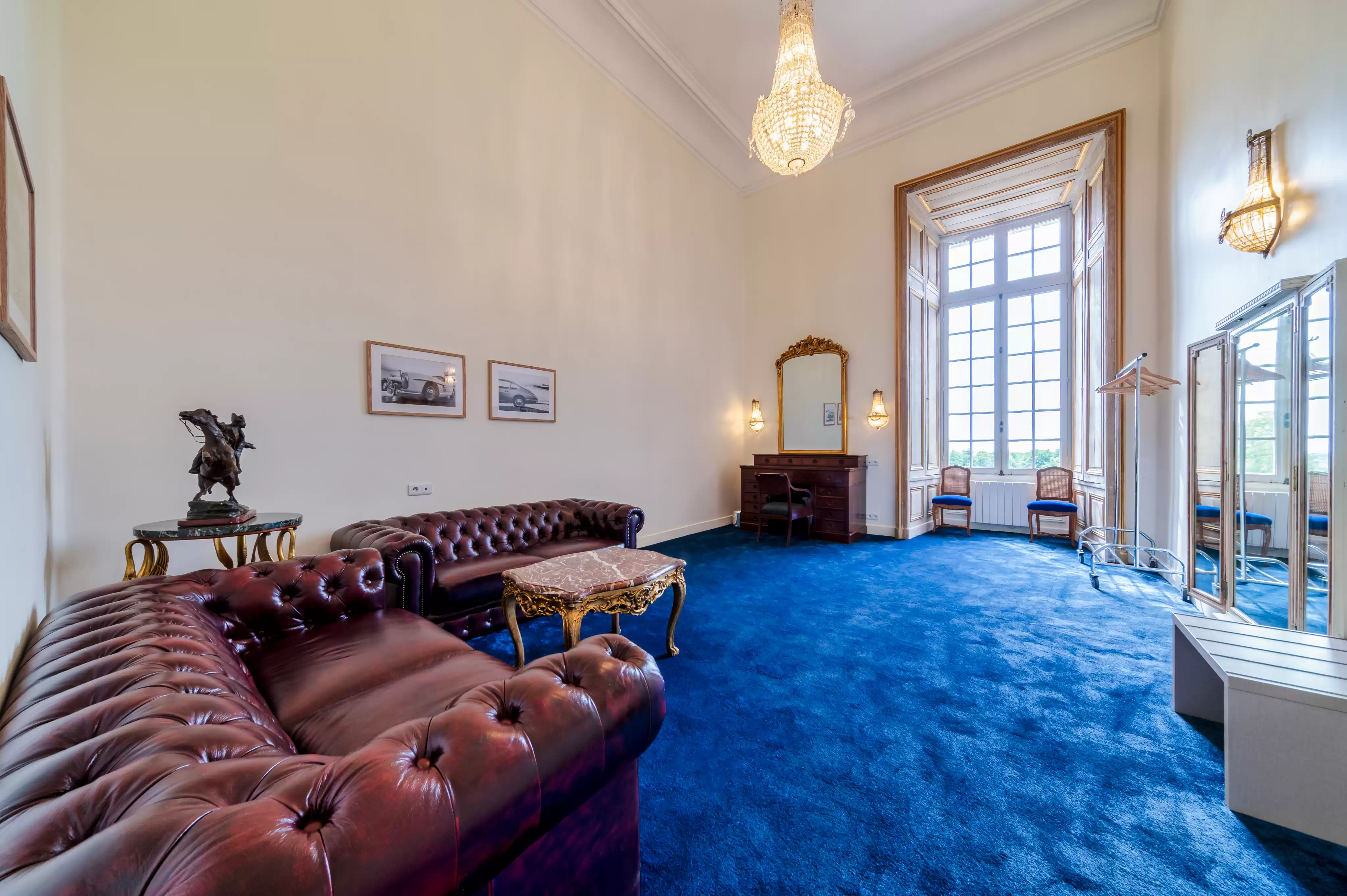 Salon de préparation du marié spacieux et luxueux, aménagé avec un canapé Chesterfield en cuir marron, un bureau de style ministre, et une moquette bleu roi sous un lustre en cri