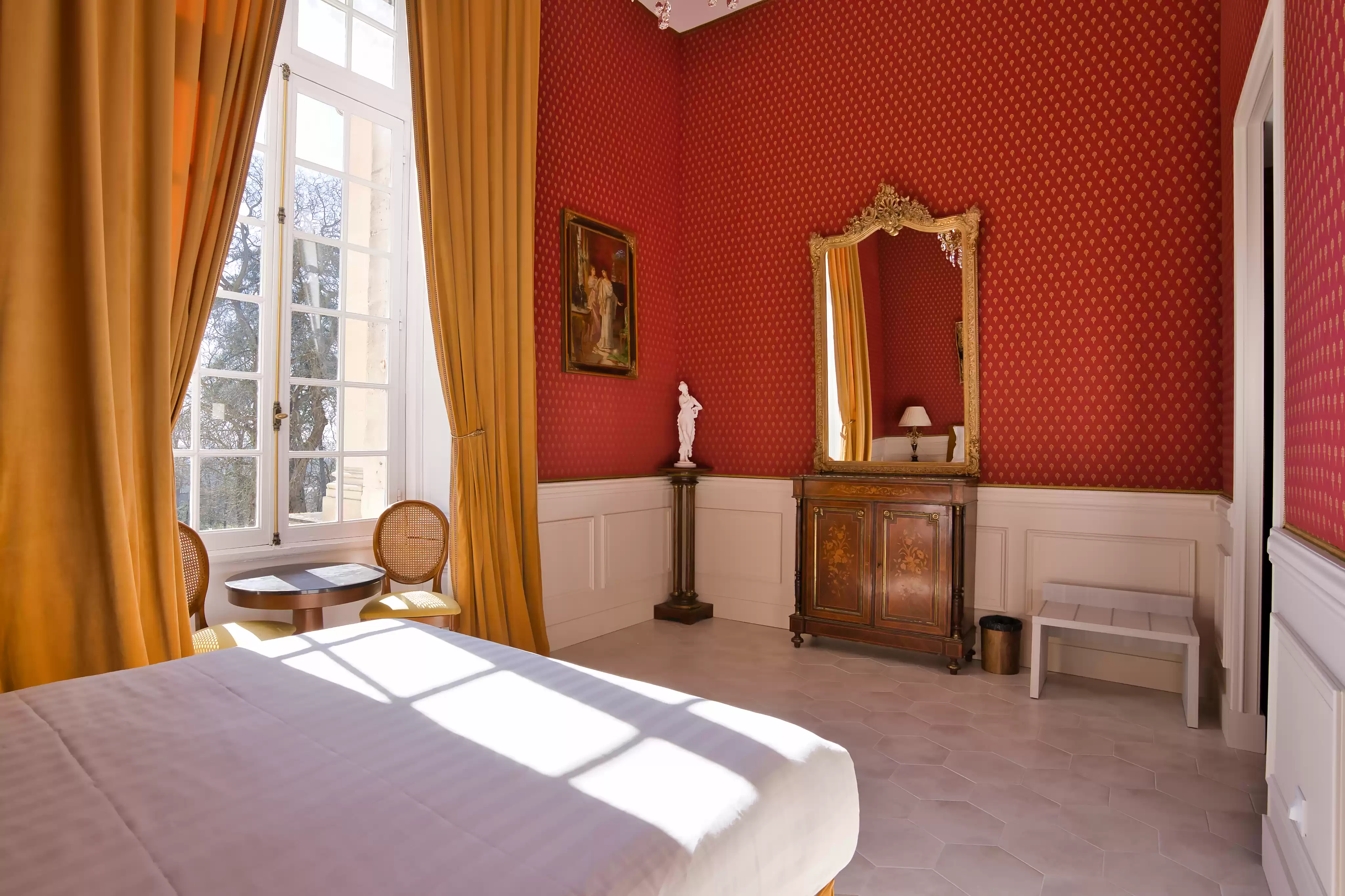 Chambre de château élégante offrant un havre de quiétude, avec des murs rouges damassés, de grands rideaux dorés, la lumière du soleil sur le lit, et un mobilier d'époque 