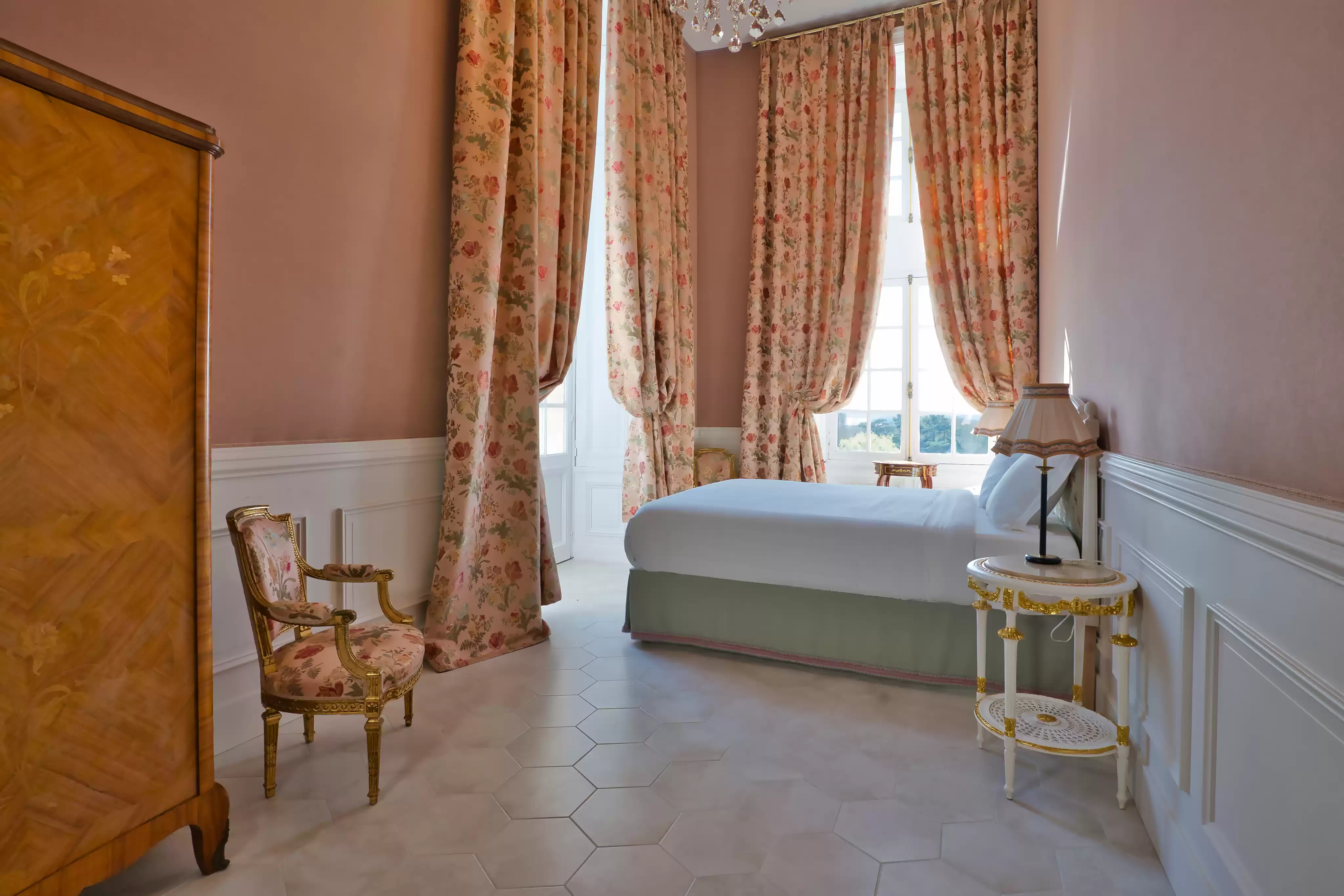 Chambre à l'atmosphère douce et lumineuse, décorée de murs roses, de longs rideaux à motifs floraux, d'un fauteuil d'époque et d'une armoire en bois 